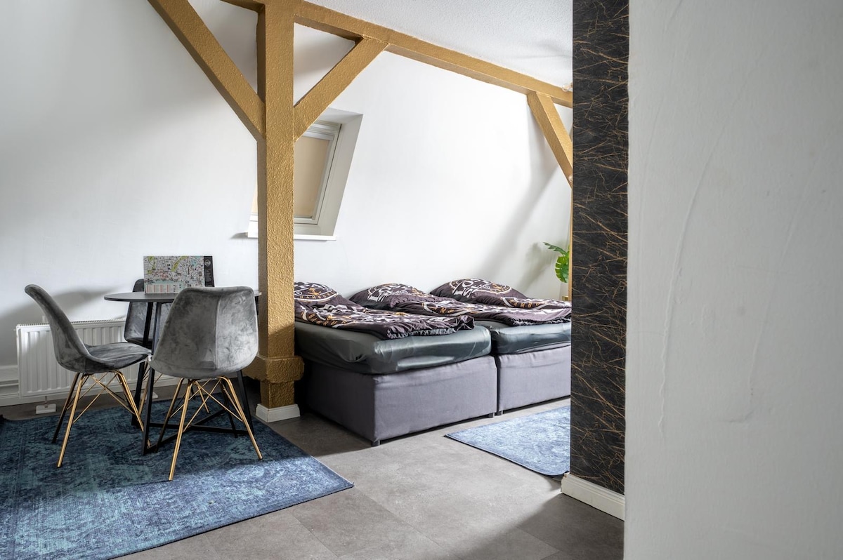 A cozy living area is presented, featuring a dining table with modern chairs and two large beds covered in patterned bedding. Soft area rugs are placed on the floor, with natural light coming through a window set into the sloped ceiling.