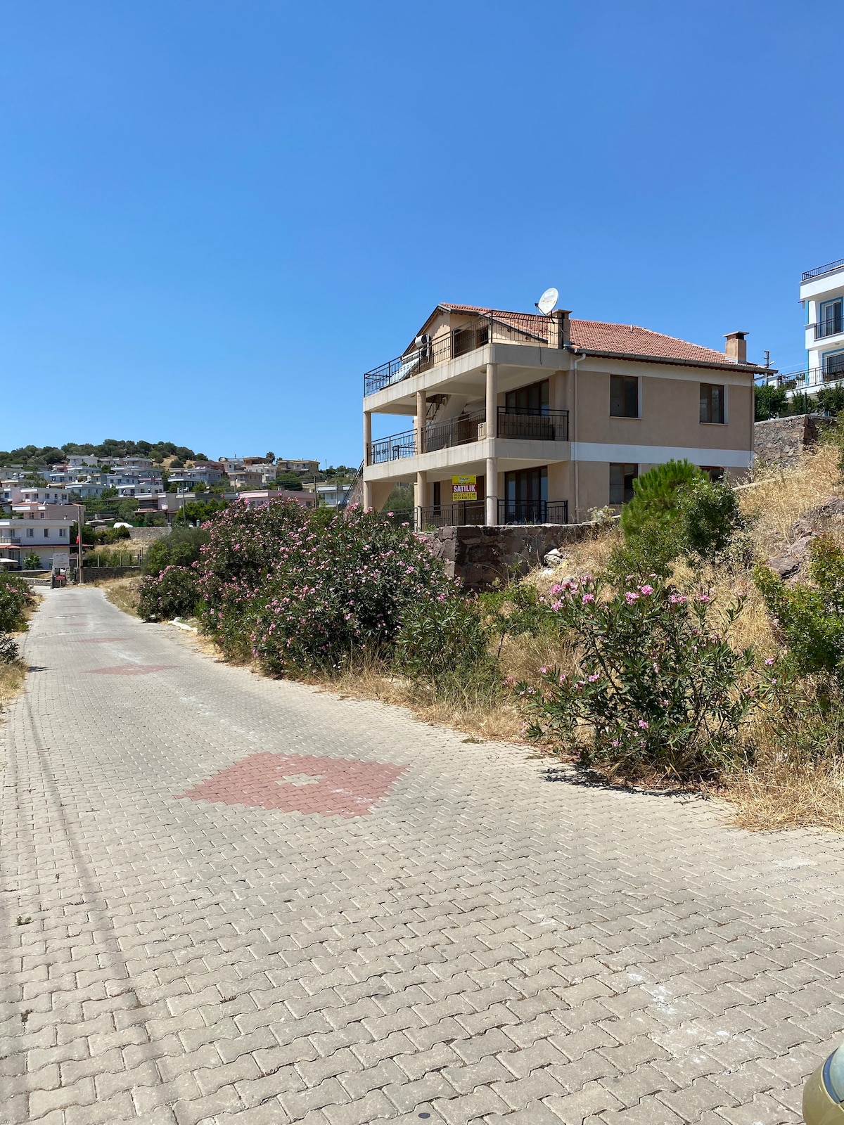 A modern, two-story building is seen with a red tile roof and balconies overlooking the surroundings. Stone pathways lead to the property, bordered by lush vegetation and blooming flowers. The clear blue sky provides a bright backdrop for the tranquil setting.