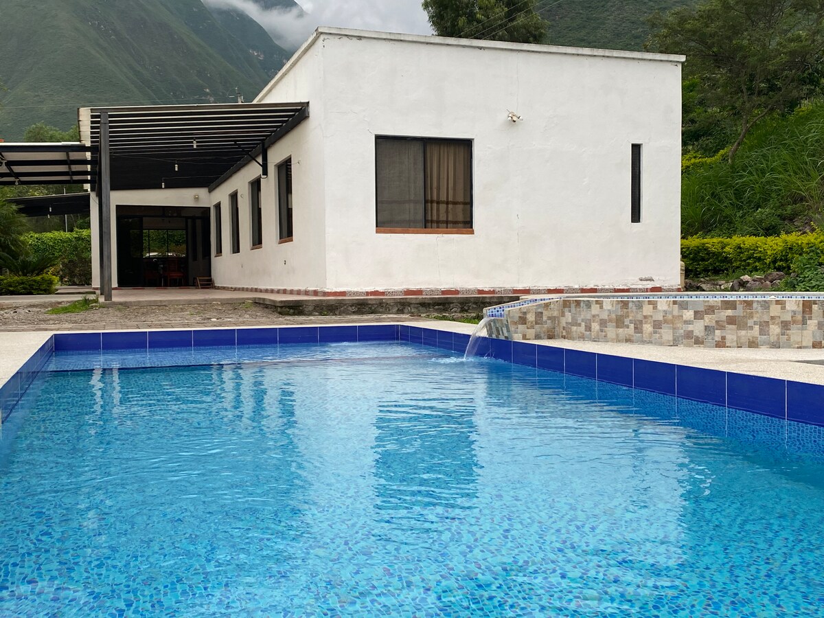 A modern house is positioned next to a clear swimming pool, reflecting the sky and surrounding greenery. The pool's edge features a small waterfall that adds a tranquil sound. Large windows are visible, allowing natural light to brighten the interior spaces.