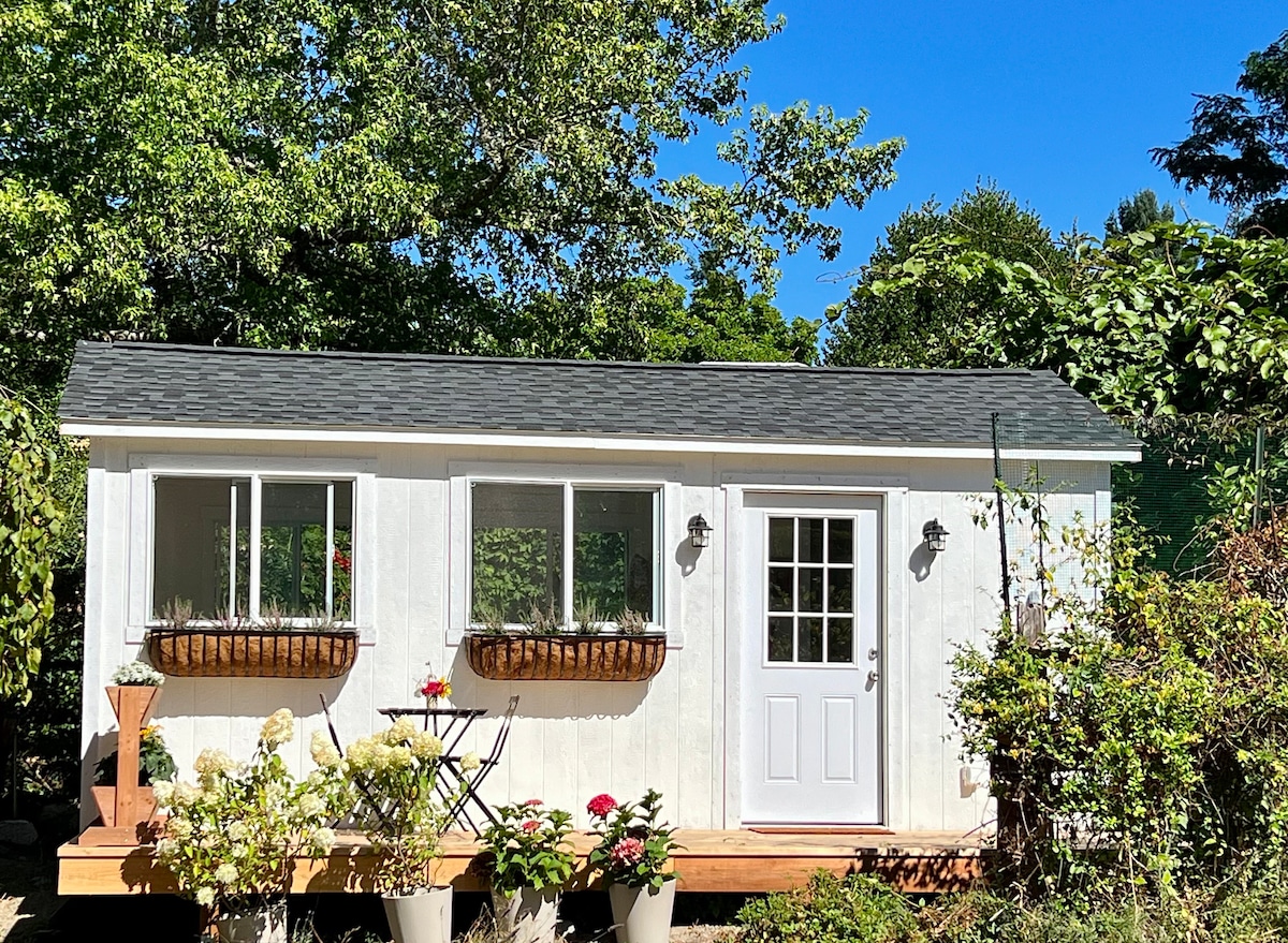 A cozy tiny cottage is framed by lush green trees and vibrant plant life. The structure features large windows adorned with flower boxes, and a welcoming front door leads to a spacious deck, perfect for outdoor relaxation.