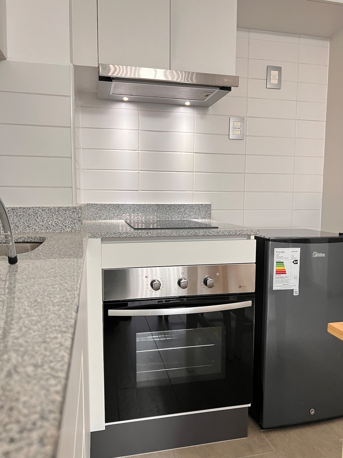 The modern kitchen features sleek cabinetry, a built-in oven with a cooktop, and a minimalist gray countertop. Under-cabinet lighting illuminates the workspace, while a compact refrigerator is positioned to the right, offering essential appliances for a convenient stay.