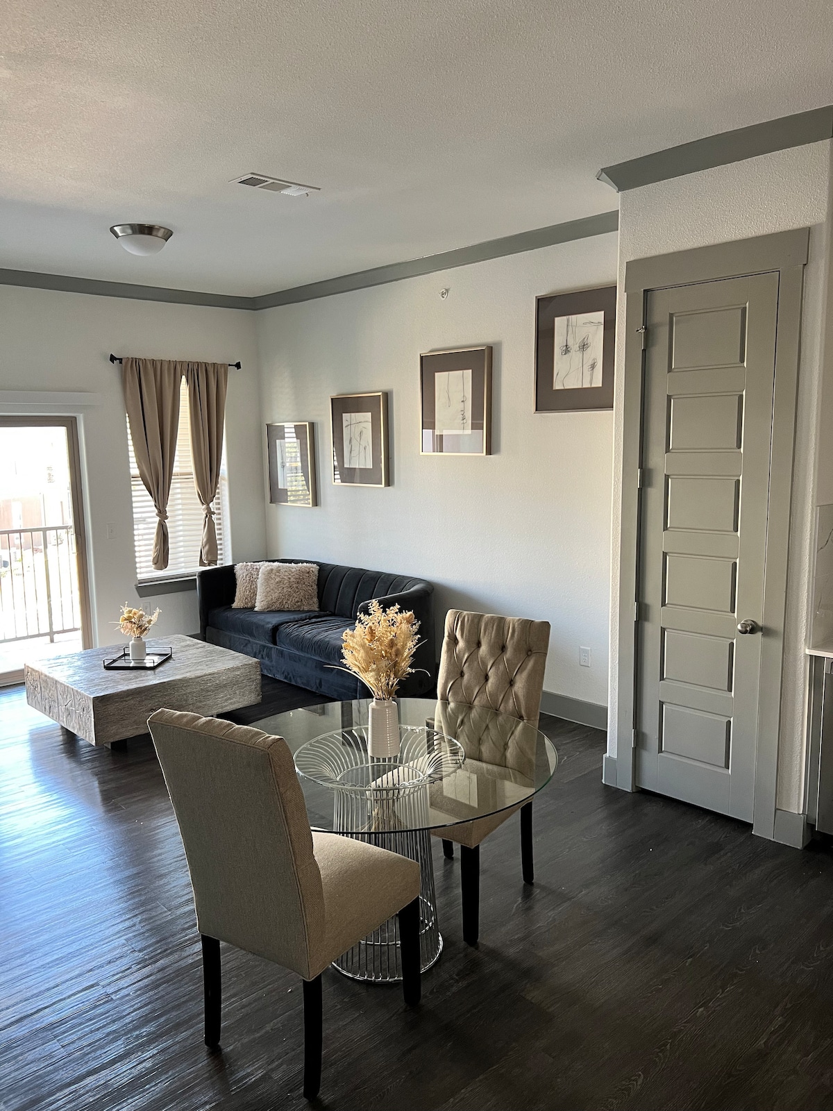 A stylish living area is highlighted by a dark sofa positioned against a light wall. A glass-top table is surrounded by four upholstered chairs. Framed artwork decorates the walls, and natural light filters in through the window, creating a welcoming ambiance.
