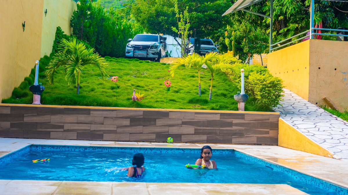 A serene outdoor space features a pool surrounded by lush greenery. Two individuals are seen enjoying the water. In the background, vehicles are parked on a grassy area, with a sloped path leading to the entrance.