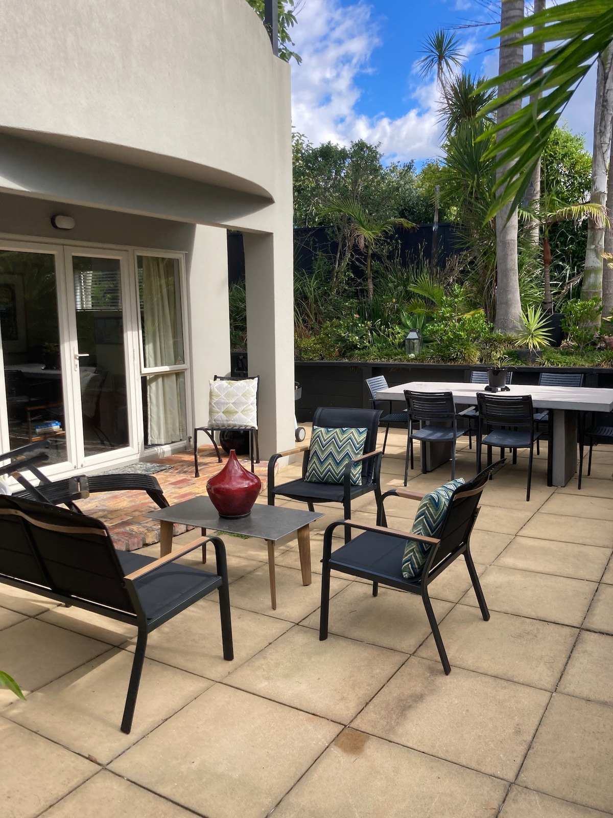 A spacious outdoor courtyard features a mix of seating arrangements, including four black chairs with patterned cushions around a low table. A dining area with a large table is also visible. Lush greenery and palm trees provide a private and relaxing environment.