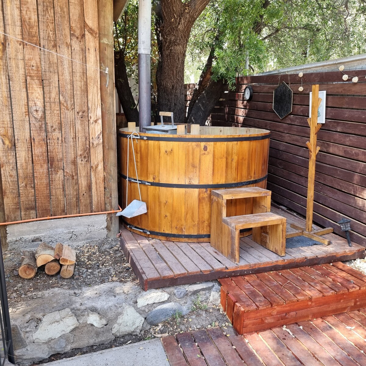 A wooden hot tub is nestled next to a rustic cabin, surrounded by natural elements. Sturdy wooden steps lead up to the tub, with a backdrop of a large tree and a wooden fence providing privacy. Piled logs are arranged nearby.