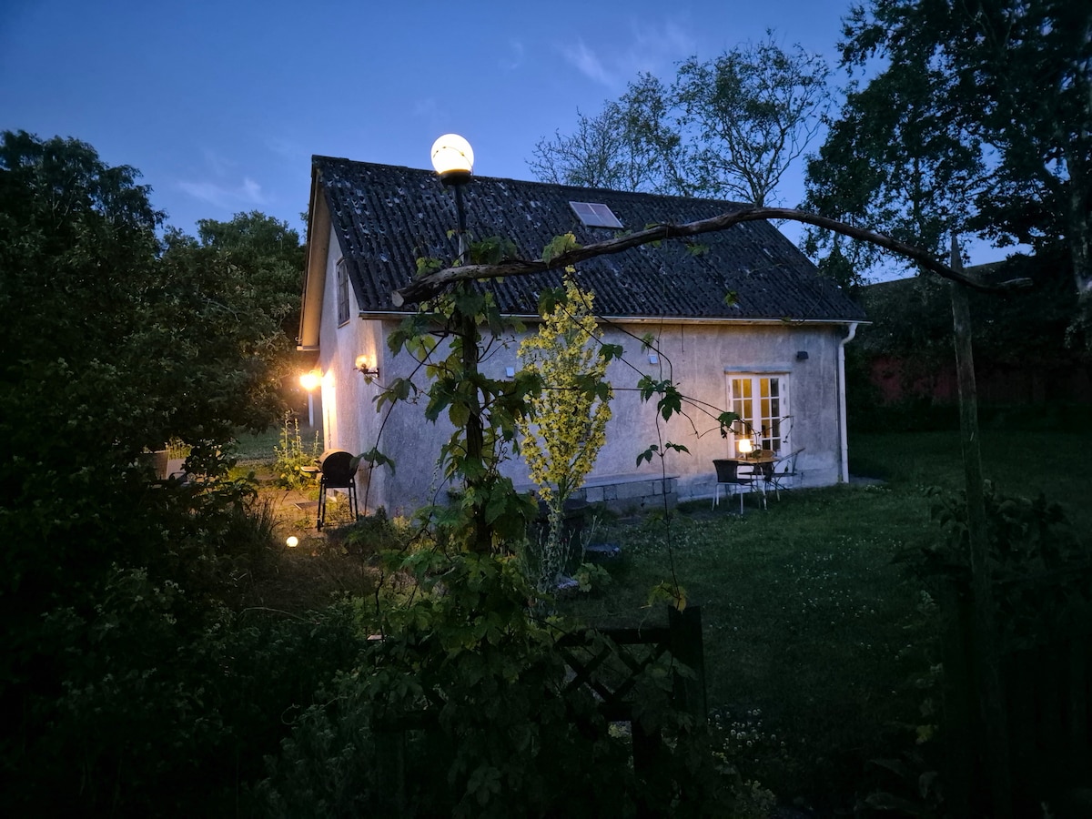 The exterior of a charming two-story house is illuminated by soft garden lights during twilight. A small table and chairs are visible on the porch, nestled among the greenery, while trees surround the property, creating a peaceful ambiance.