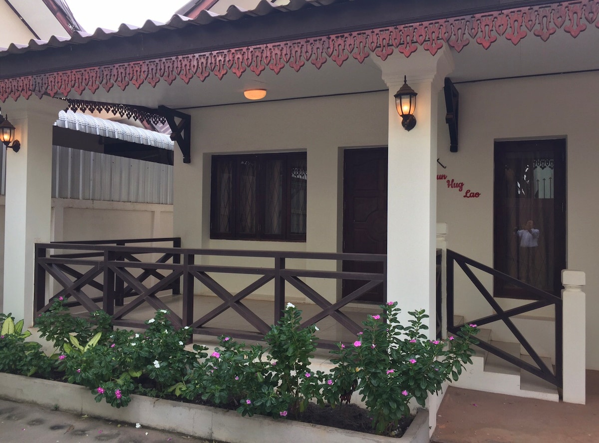 A charming exterior of the house features a covered porch with decorative wooden railings and two lantern-style lights. The path is bordered by green plants and flowers. Windows are adorned with drapes, providing a welcoming entrance to the home.