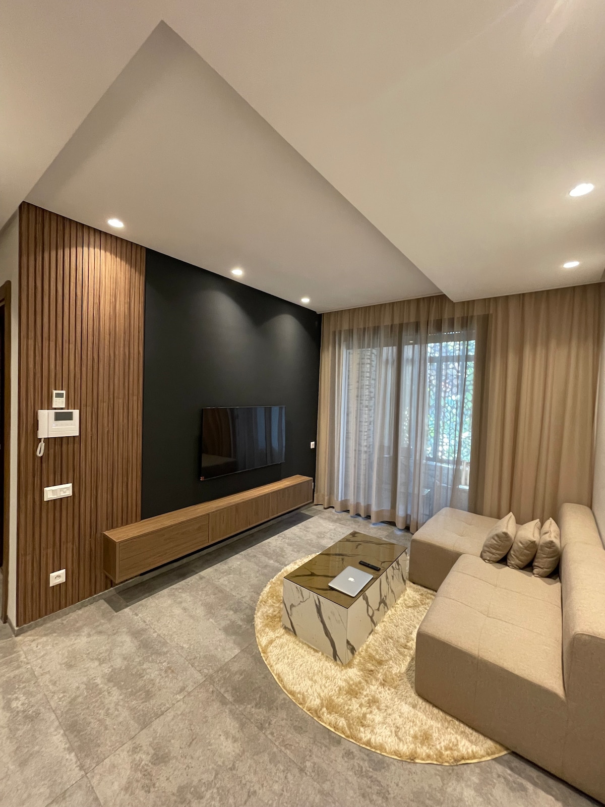 A modern living area features a light-colored sofa arranged around a stylish coffee table on a circular rug. A flat-screen TV is mounted on the wall in front, with warm lighting fixtures illuminating the space. Sheer curtains frame a window, allowing soft natural light to enter.
