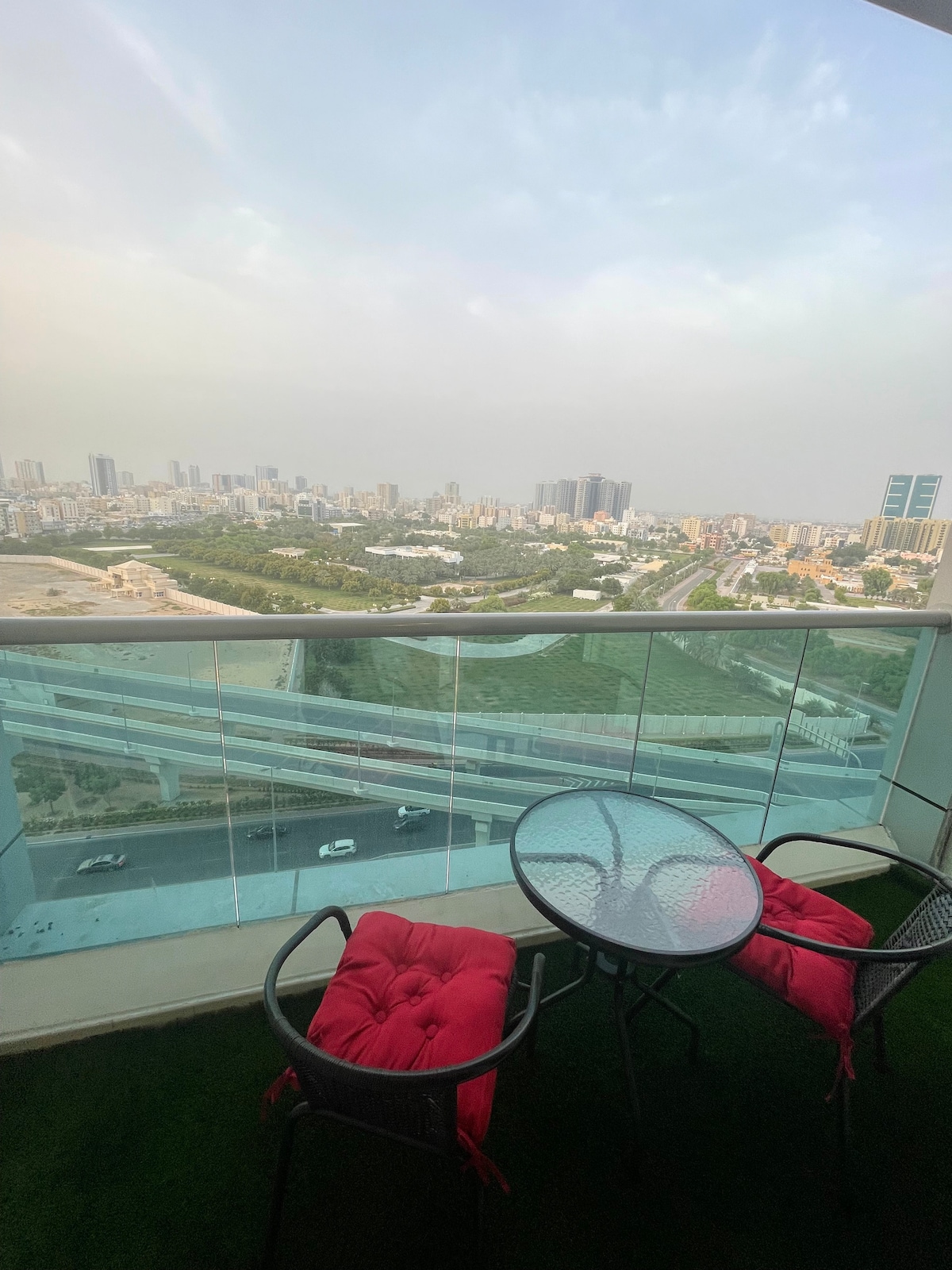 A glass-enclosed balcony offers a view of the city skyline and surrounding greenery. A round glass table is paired with two black chairs, featuring soft red cushions. The flooring is covered with artificial grass, enhancing the outdoor experience.