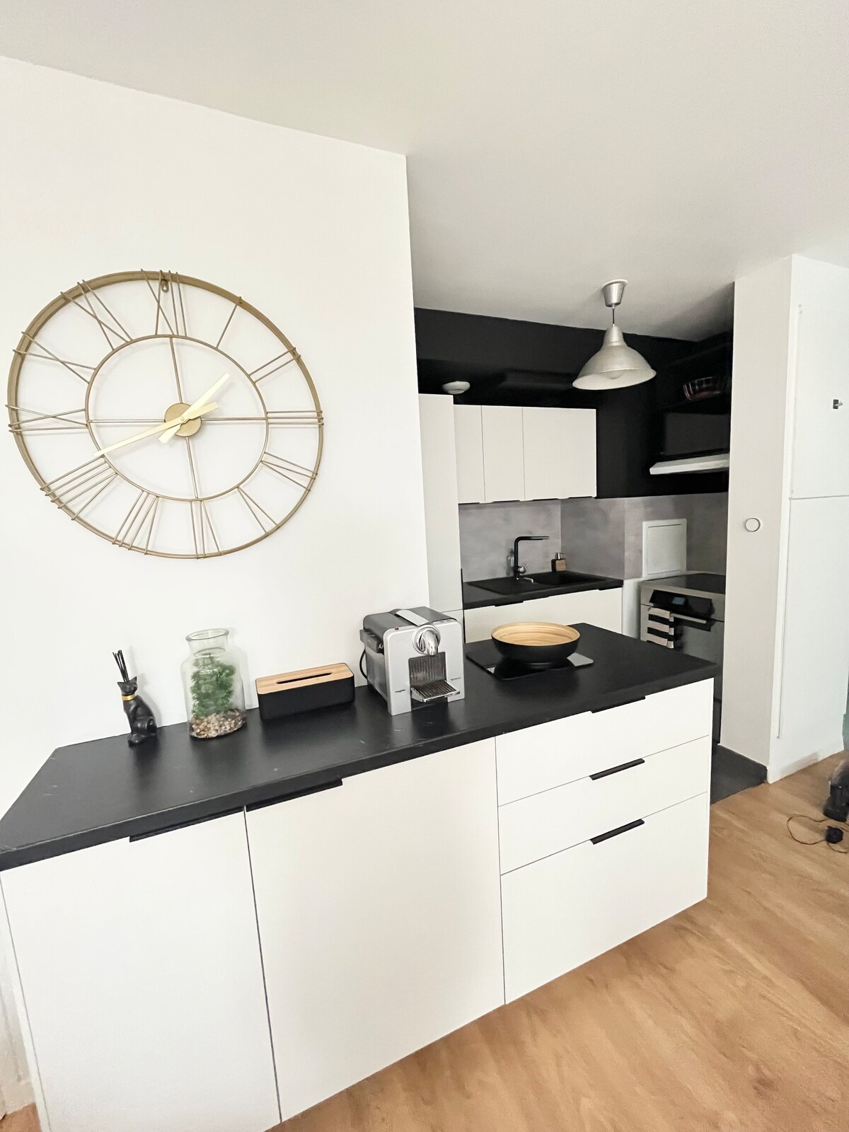 A modern kitchen area features sleek white cabinetry and a black countertop. A large wall clock adds a stylish touch, while appliances are neatly arranged. The design incorporates minimal decor, enhancing functionality and a clean aesthetic.