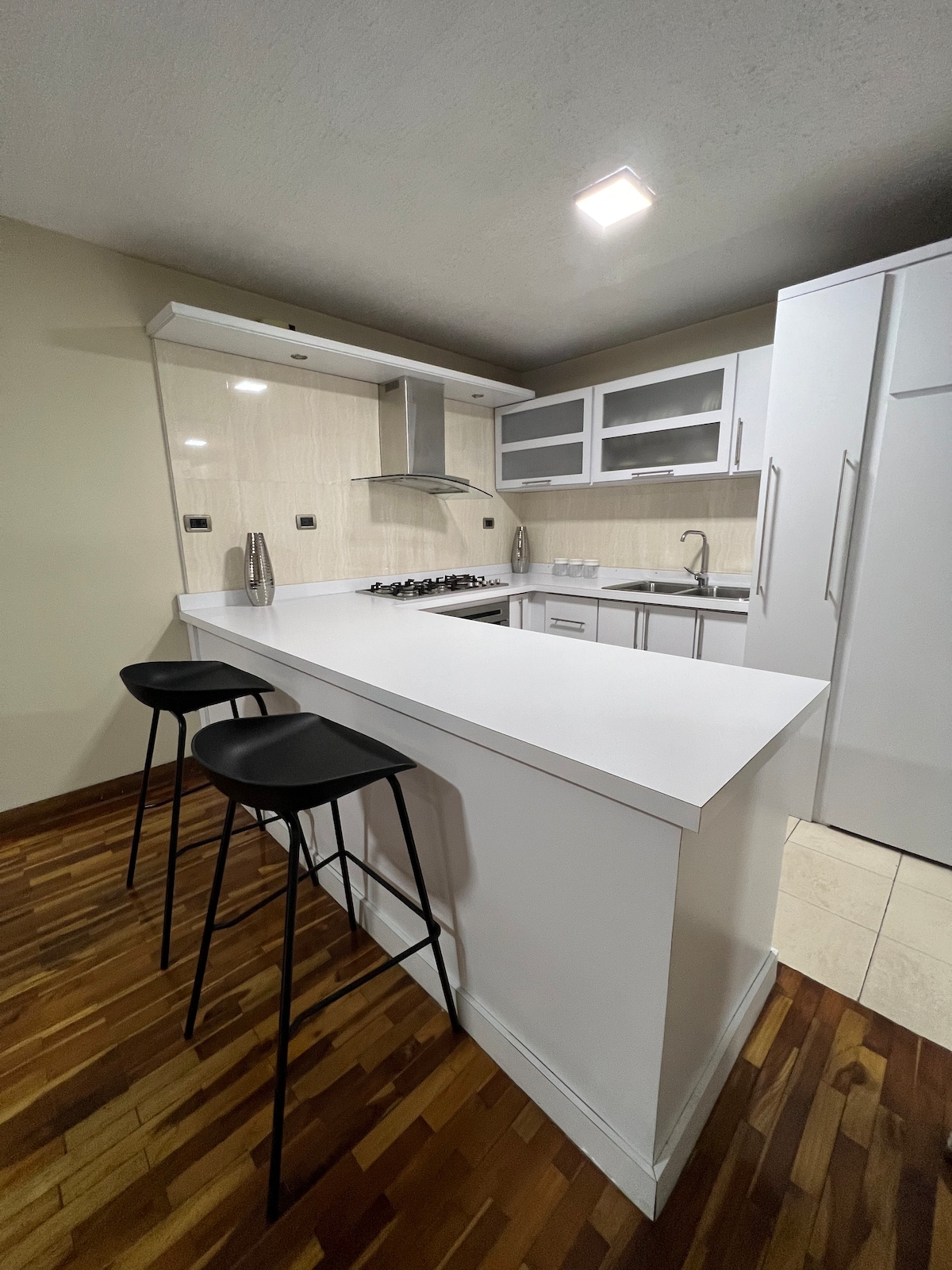 A modern kitchen space features sleek white cabinetry and an island with two high-backed black stools. Stainless steel appliances are visible, alongside a gas stove and a sink. Warm wood flooring contrasts with the light-colored surfaces, creating a clean and inviting cooking environment.