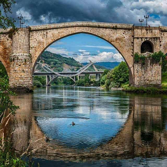 An arched stone bridge spans a calm river, reflecting its structure in the water below. Surrounding greenery complements the natural landscape, while a modern bridge in the background contrasts with the historical architecture of the bridge in the foreground.