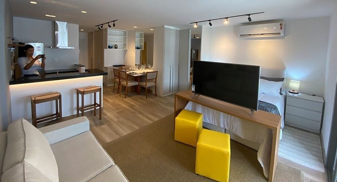 A modern living space is highlighted, featuring a cozy seating area with a light-colored sofa and two bright yellow stools. A large flat-screen TV is positioned on a wooden media console. The open kitchen and dining area are visible, showcasing sleek cabinetry and a well-set table.
