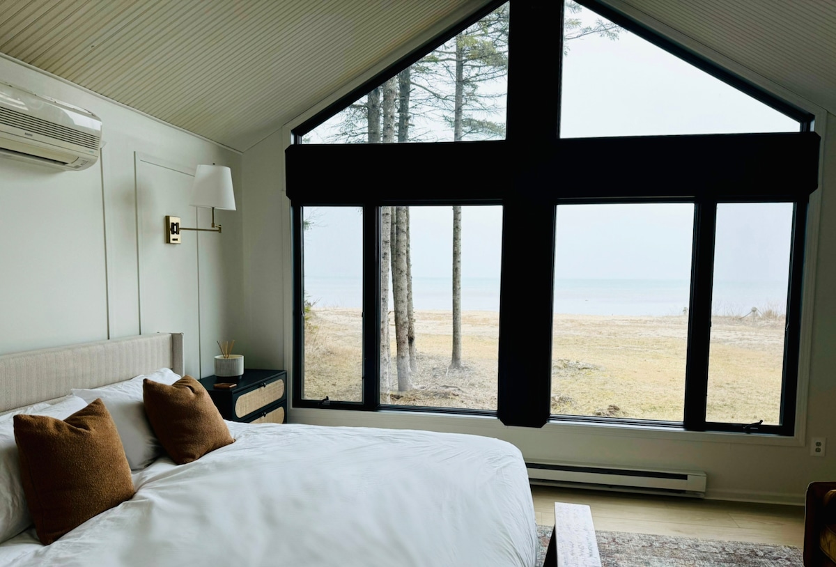 A spacious bedroom features a large window with black coverings, offering views of the adjacent sandy beach and Lake Michigan. The room includes a comfortable bed adorned with plush pillows, while neutral tones create a calm and serene environment.