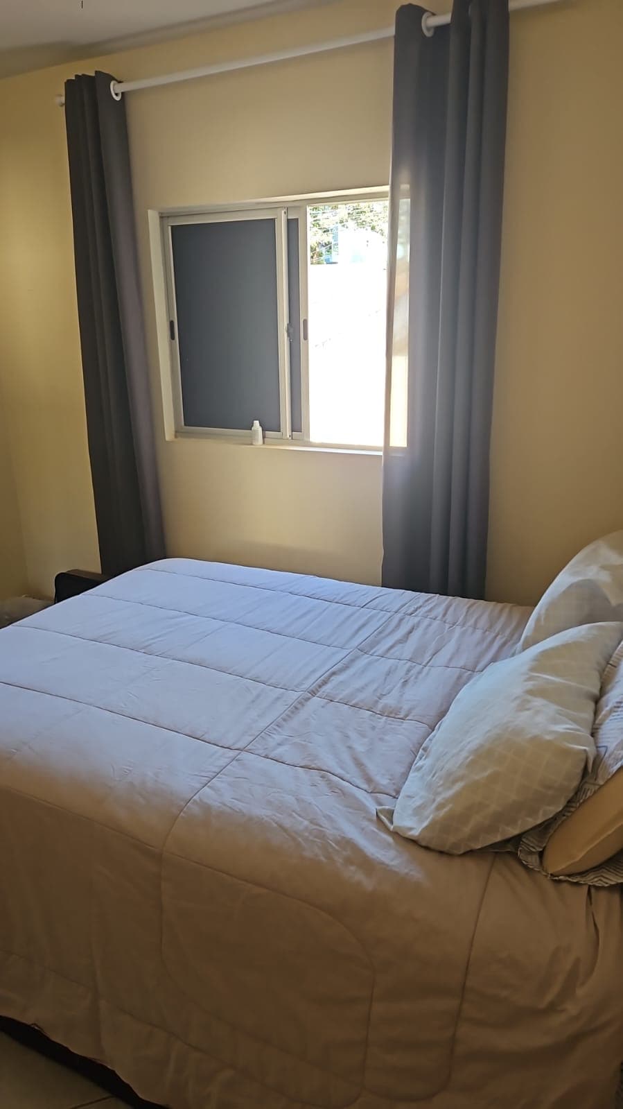 A room featuring a spacious bed covered with light bedding and accented by a decorative pillow. Natural light filters through a nearby window, framed by partial blackout curtains, creating a soft ambiance in the room.