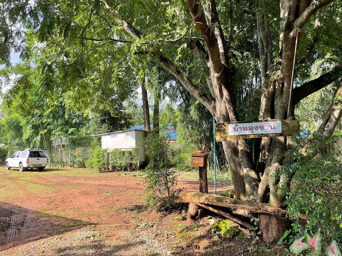 A welcoming entrance is marked by a sturdy wooden sign hanging from a large tree, indicating the property name. A grassy area surrounds a well-maintained dirt path leading to a small house. A parked vehicle and lush greenery provide a serene rural backdrop.