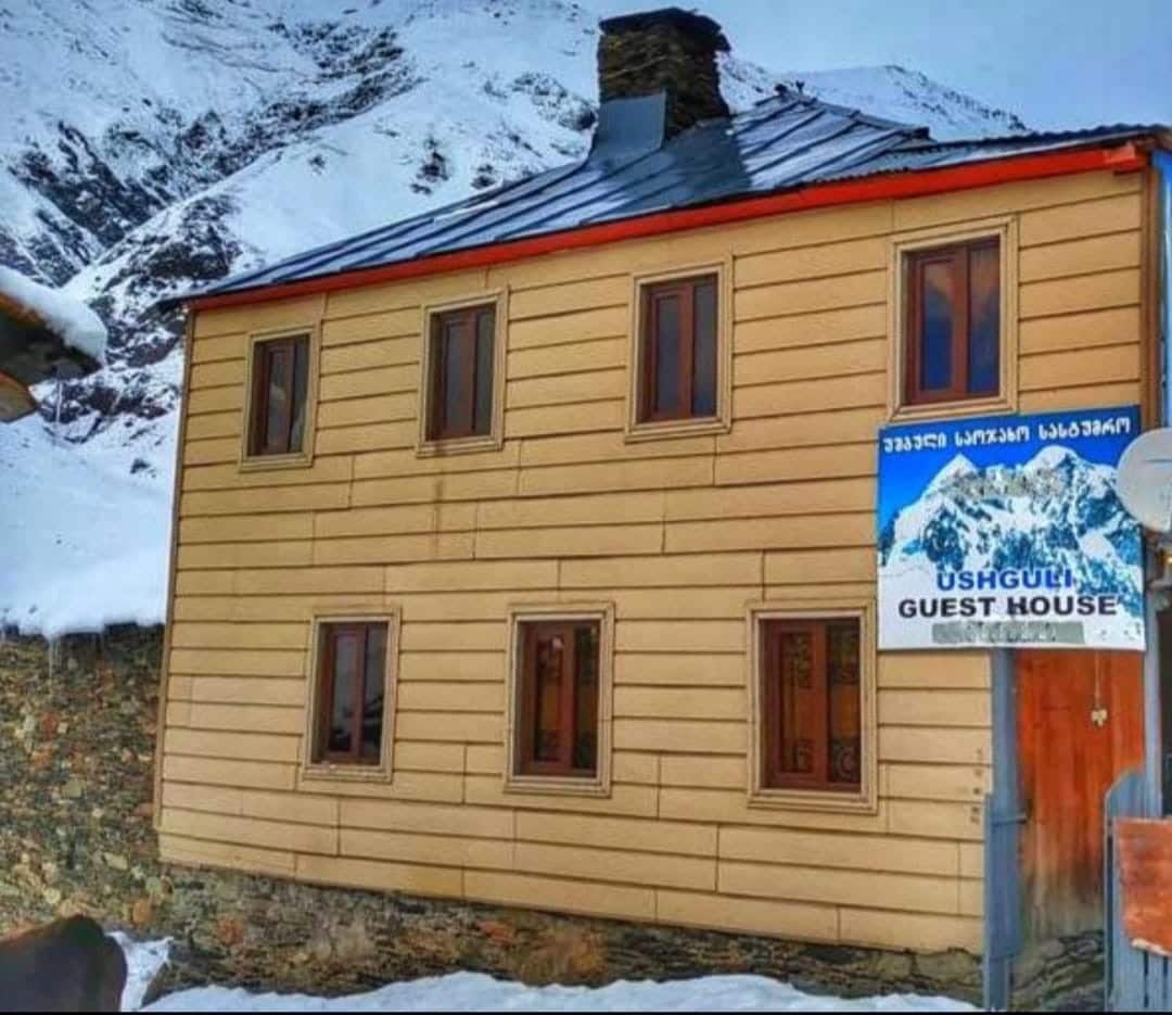 The exterior of Guest House Buba is shown, featuring a two-story structure with a smooth wooden facade. Snow covers the ground, and several windows are visible, framed in red. A sign indicating the guest house is positioned nearby, with a backdrop of rocky mountains in the distance.