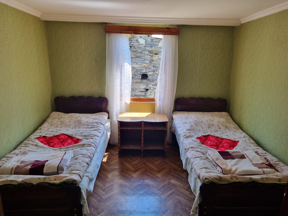 A guest room is presented with two beds, each dressed in floral-patterned linens. Natural light enters through a window adorned with sheer curtains, illuminating the wooden furniture, including a small table positioned between the beds. The walls are painted in a soothing green hue.