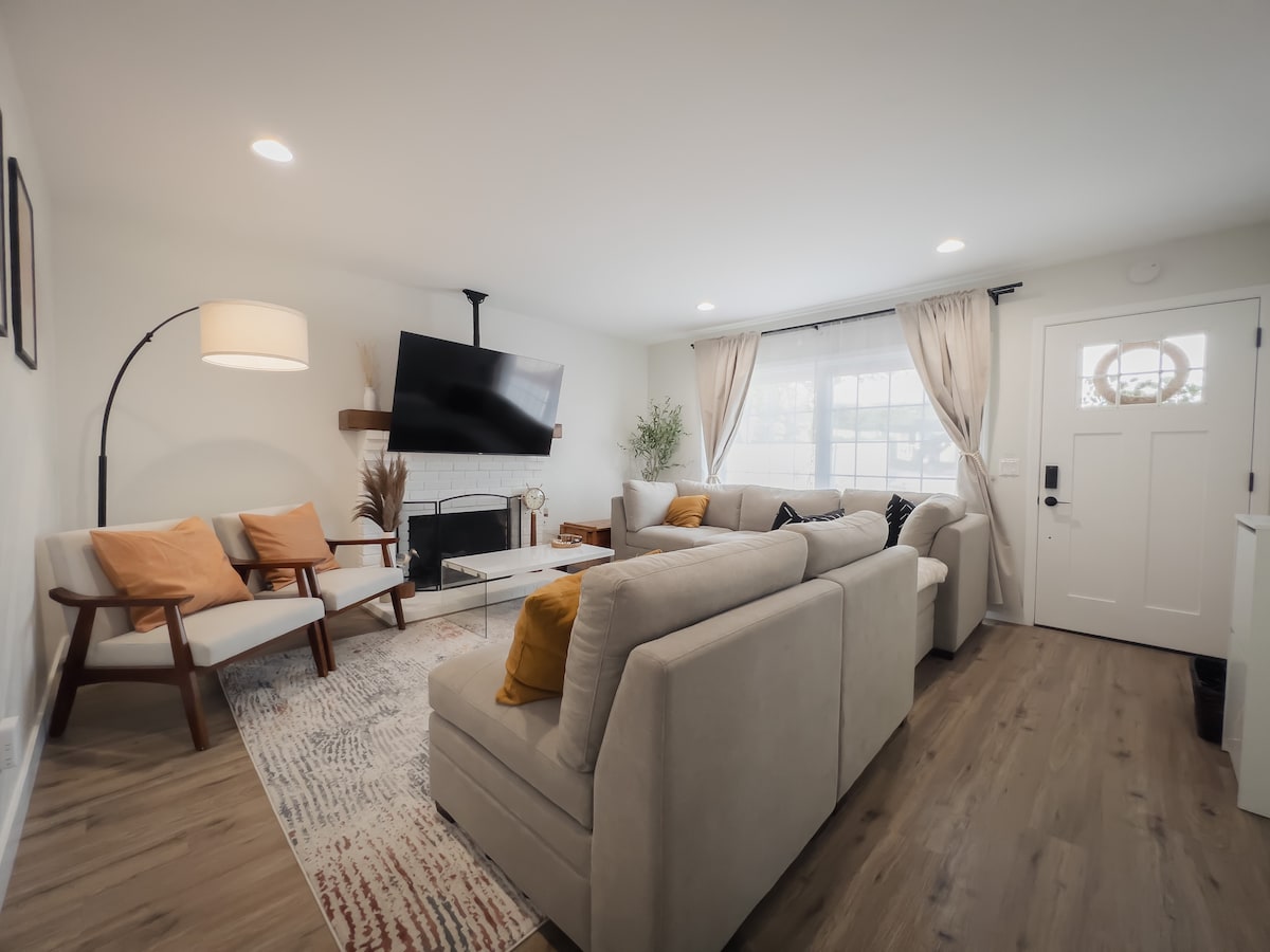 A modern living area is appointed with a spacious sectional sofa and accent chairs. Soft neutral colors are complemented by warm throw pillows. A flat-screen TV is mounted on the wall, while natural light filters through the large window, enhancing the cozy ambiance.