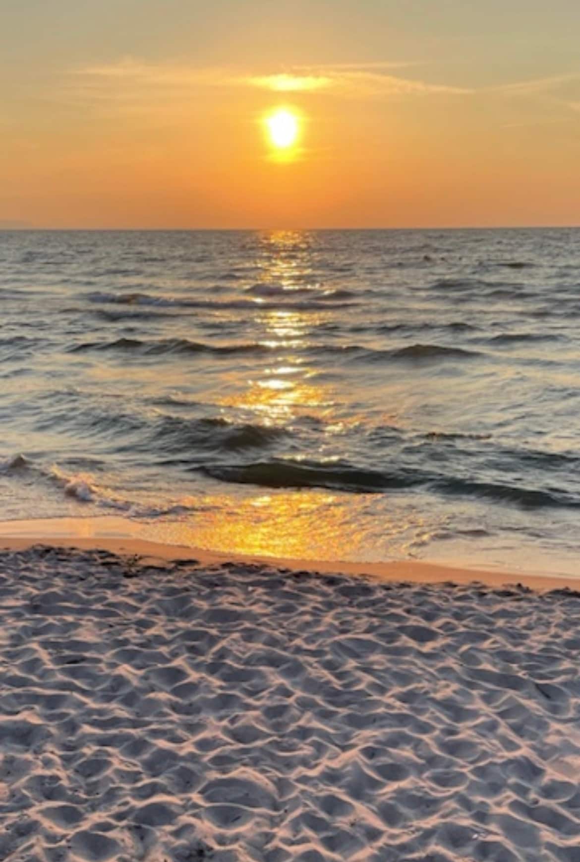 A tranquil beach scene at sunset is captured, displaying soft waves gently lapping against the shore. The sky is painted in warm tones of orange and gold, reflecting on the water's surface. The sandy foreground creates a serene transition to the calm sea.