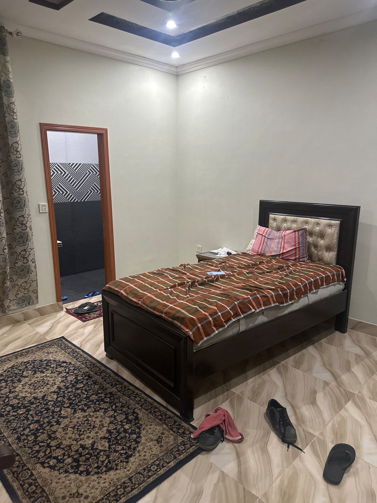 A simple bedroom is depicted, featuring a bed with a patterned blanket. A dark wooden bed frame is complemented by a headboard. The room's light-colored walls contrast with a dark rug on the floor, while some footwear is scattered nearby.