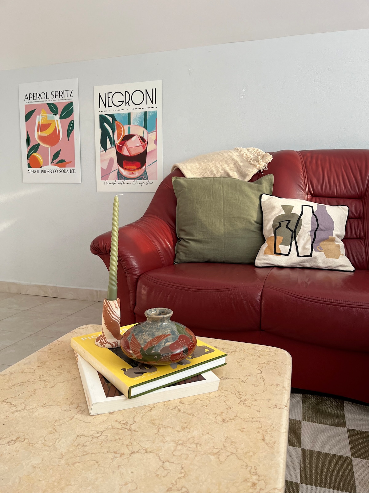 A comfortable red sofa is adorned with decorative cushions, including a green one and a patterned one. A marble coffee table displays a stack of books, a unique vase, and a candle. Art prints featuring cocktail illustrations are hung on the wall.
