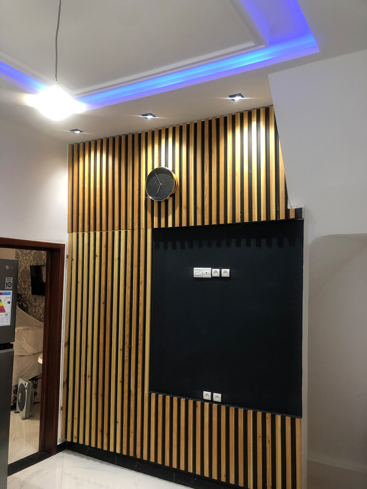 A wall features a modern design with vertical wooden slats and a round wall clock positioned near the top. Below, a chalkboard area is outlined, complemented by light switches. Soft blue lighting from the ceiling adds a calming touch to the contemporary space.