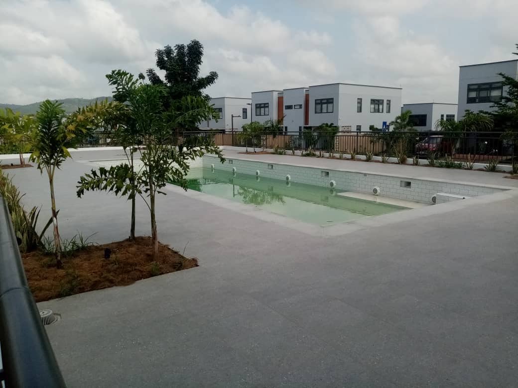 An expansive outdoor area features a calm swimming pool surrounded by greenery. Modern townhouses are visible in the background, set against a partly cloudy sky. The pool area includes subtle lighting and is bordered by neatly arranged landscaping.