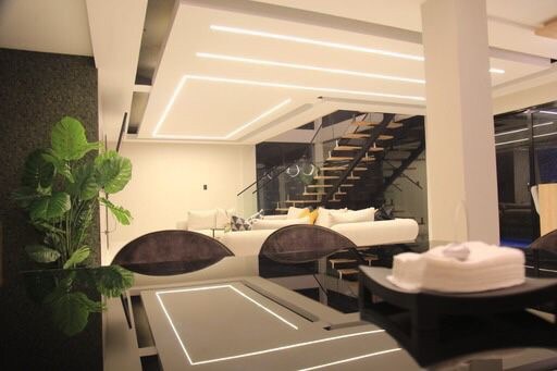 A modern living space is depicted, featuring a spacious sofa with light-colored cushions and a sleek staircase leading to the upper level. Green plants add a touch of nature, while recessed lighting creates a soft glow throughout the open area.