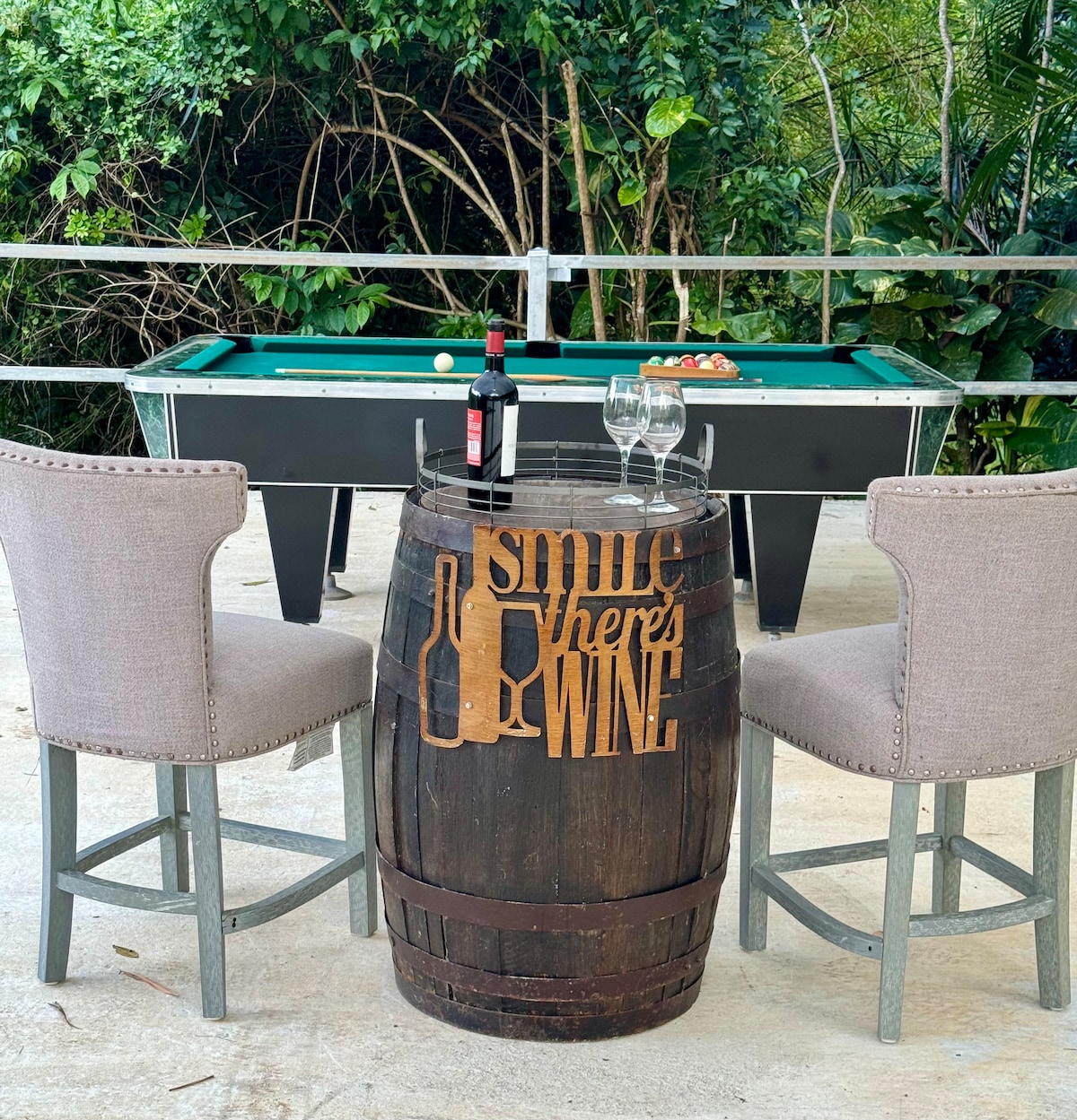 A wooden barrel table with an engraved design sits surrounded by two light-colored chairs. A pool table is visible in the background, adding a recreational element to the space. Lush greenery provides a natural backdrop, enhancing the inviting atmosphere.