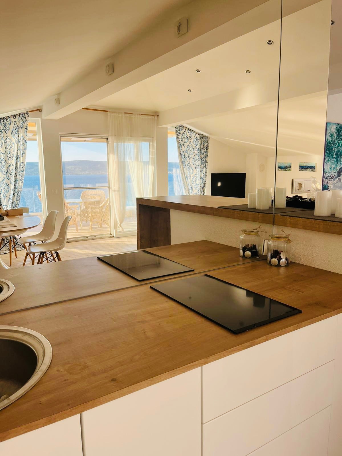 The modern kitchen features a wooden countertop with sleek black cooktops, reflecting a contemporary design. A large window offers a view of the sea, while light enters the space through bright curtains. Dining space is visible in the background, enhancing the open atmosphere.