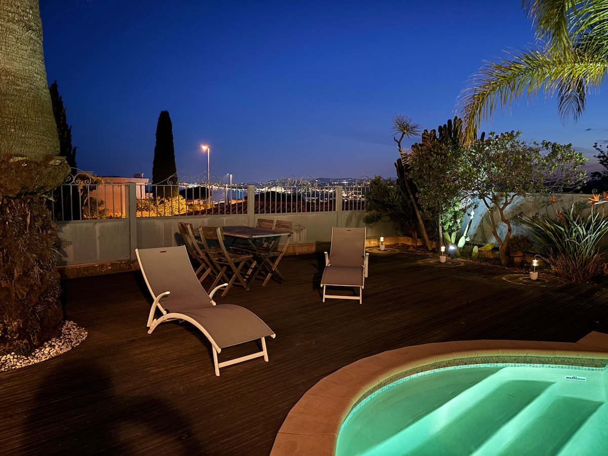An outdoor area is illuminated at night, featuring a small pool with inviting turquoise water. Sun loungers are positioned around the pool, and a wooden table and chairs provide a dining space. Lush greenery and city lights create a serene atmosphere.