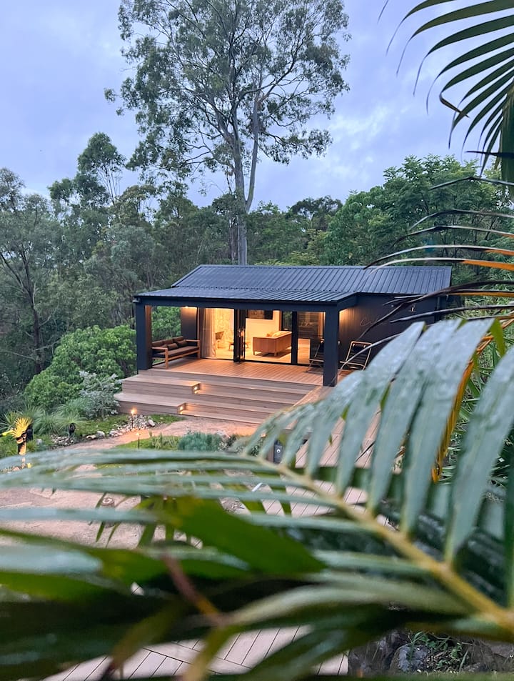 A cozy, modern cabin with an illuminated wooden deck sits nestled in a lush, forested hillside. The cabin has an inviting open front with warm lighting inside, and steps leading down to a landscaped garden. Palm leaves frame the foreground, adding to the tropical, secluded feel