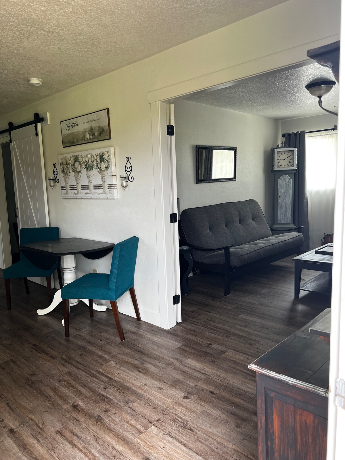 A cozy entryway is shown, featuring a small table with two turquoise chairs and a gray futon in the adjacent living area. Natural light enters through a window covered with light curtains, illuminating the wood-like flooring and simple décor.