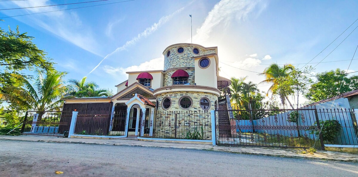 A unique two-story house is framed by palm trees and surrounded by a gated perimeter. The exterior features a combination of stone and stucco with circular windows, a decorative balcony, and a bright sky overhead.