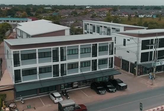 An aerial view captures a modern, multi-story building complex featuring a mix of residential and commercial spaces. Large windows provide natural light along the facade. Several parked cars are visible in the foreground, with a tree-lined street running alongside the property.