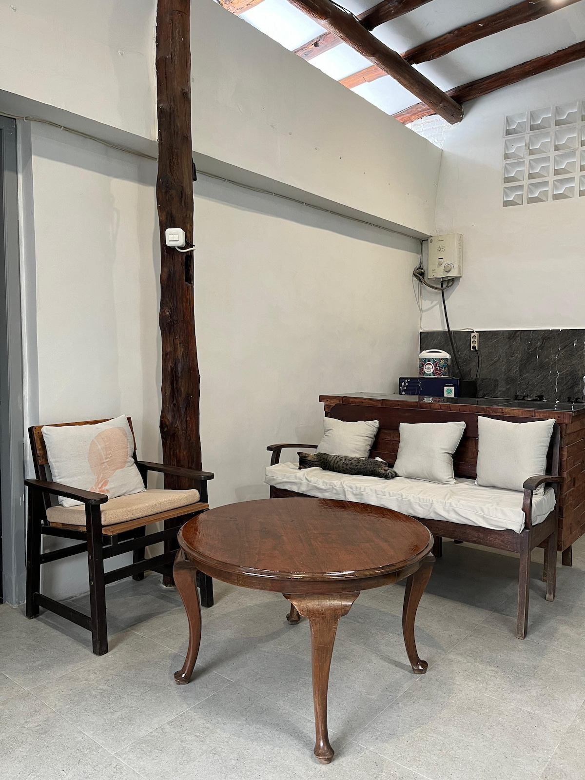 A cozy seating area is depicted, featuring a wooden table and two chairs. One chair has a comfortable cushion, while a sofa adorned with multiple pillow accents invites relaxation. The space is illuminated by natural light streaming from the ceiling.