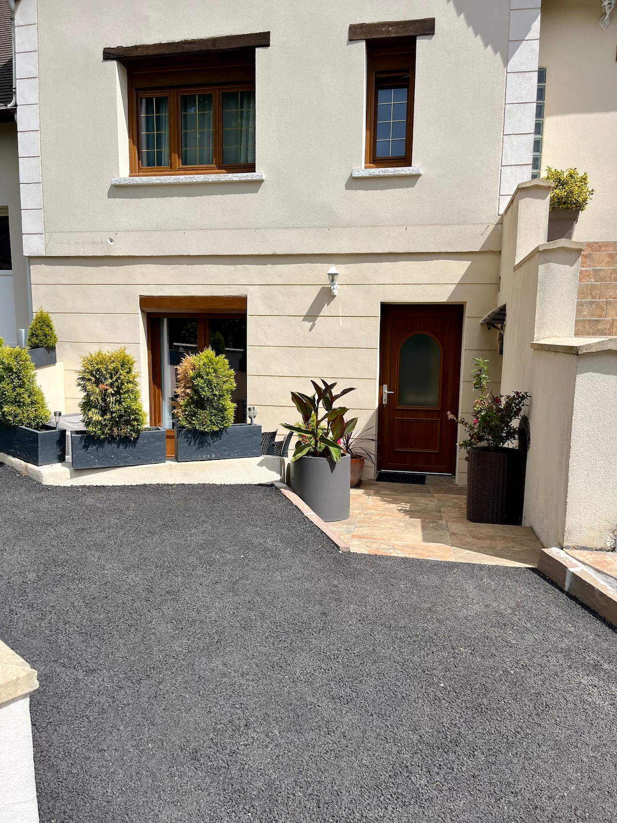 An entrance area features a welcoming door framed by neatly arranged plants in modern planters. The driveway is paved with a combination of stone and gravel, leading to the independent entrance, enhancing accessibility. The exterior walls are neutral-toned, contributing to a clean appearance.