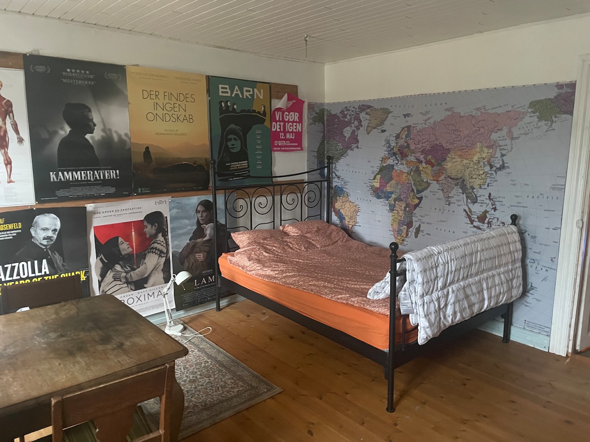 A cozy bedroom features a spacious bed with orange bedding and a white throw blanket. The walls are adorned with various posters and a large map of the world. A wooden table is set in the room, with a light-toned rug placed beneath.
