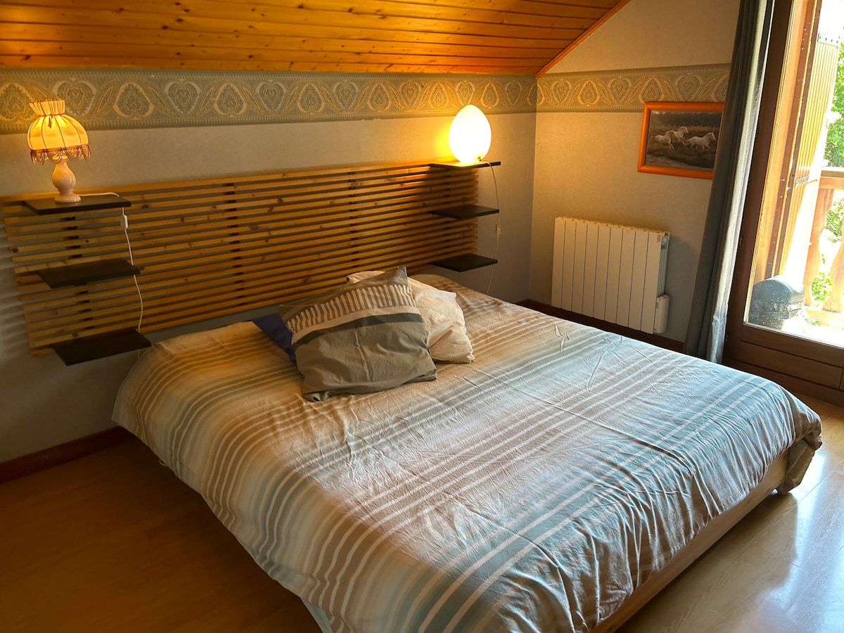A spacious bed is centered in the room, adorned with a striped blanket and several decorative pillows. A wooden headboard features built-in shelves on either side. Light is softly emitted from a bedside lamp, while natural light filters in through the window beside the bed.
