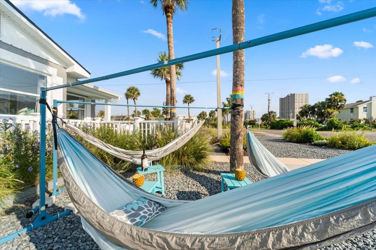 Two hammocks are suspended between tall palm trees, offering a relaxing spot to unwind. Colorful cushions rest on nearby blue stands. Lush greenery and gravel ground surround the area, all under a clear blue sky.