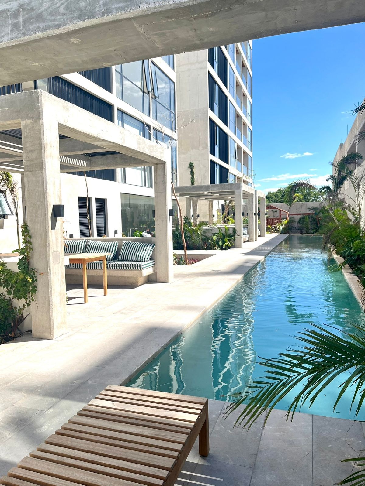 A serene outdoor area is showcased, featuring a clear swimming pool that reflects the sky. Lush greenery surrounds the space, and modern structures with concrete beams offer shade. Comfortable seating is visible beside the pool, creating an inviting atmosphere for relaxation.