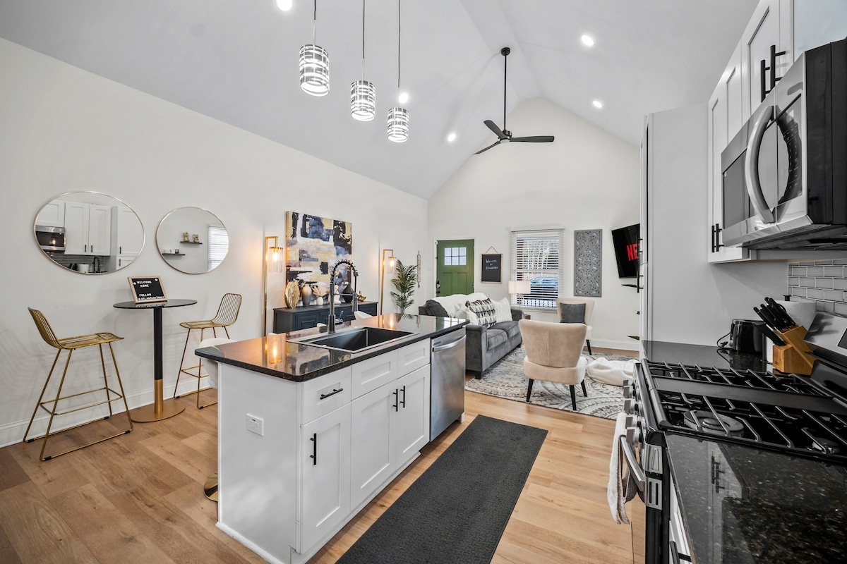 An open-concept space features high vaulted ceilings, showcasing a modern kitchen with a sleek island and top-notch appliances. Bar stools are positioned at the island, while a cozy living area is in view, enhanced by natural light from large windows.