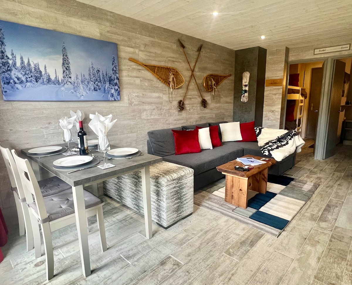 An inviting living space features a comfortable gray sofa with red and white cushions. A rustic wooden coffee table is positioned on a patterned rug, while a dining area with a gray table and chairs sits nearby. Ski decorations adorn the wall, enhancing the alpine ambiance.