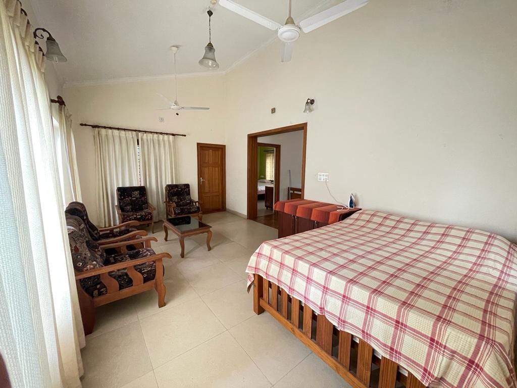 A spacious bedroom is furnished with a large bed covered in a checkered blanket. Two comfortable chairs and a coffee table are positioned nearby. Natural light enters through large windows, with light-colored curtains providing a soft backdrop.