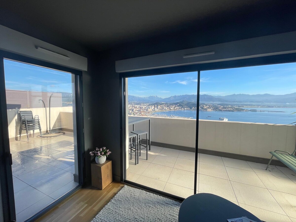 Large glass doors provide access to a spacious terrace with views of the coastline and mountains. The interior features a modern design, with a cozy seating area and a small table visible. Natural light fills the space, creating a bright and open atmosphere.
