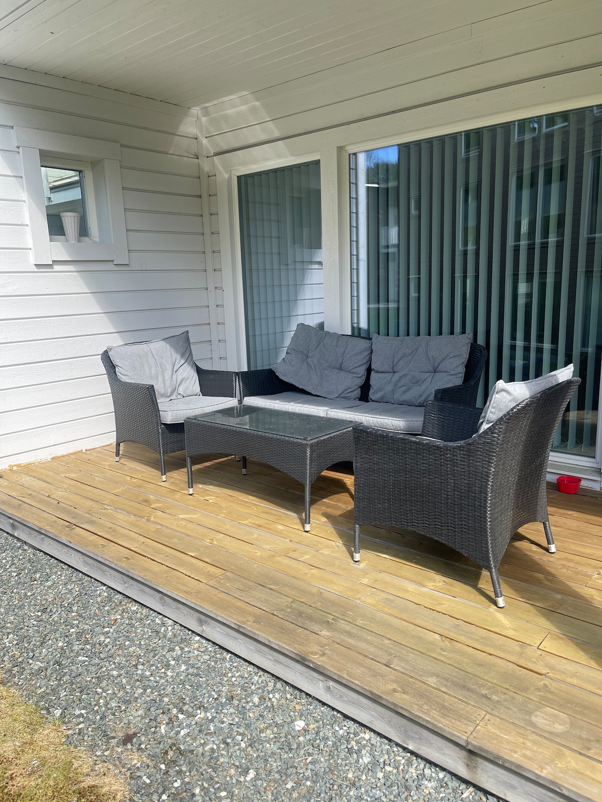 An outdoor seating area features a wicker couch with gray cushions and a glass-topped coffee table, positioned on a wooden deck. Light is filtered through vertical blinds on the window, and smooth pebbles line the ground near the seating area.