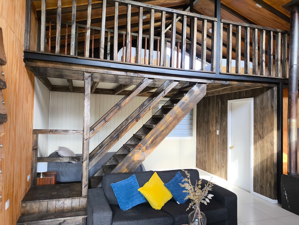 An interior space features a wooden staircase leading to a loft area with a spacious bed. A gray sofa with decorative blue and yellow cushions is positioned in front, while natural light brightens the room from a nearby window.