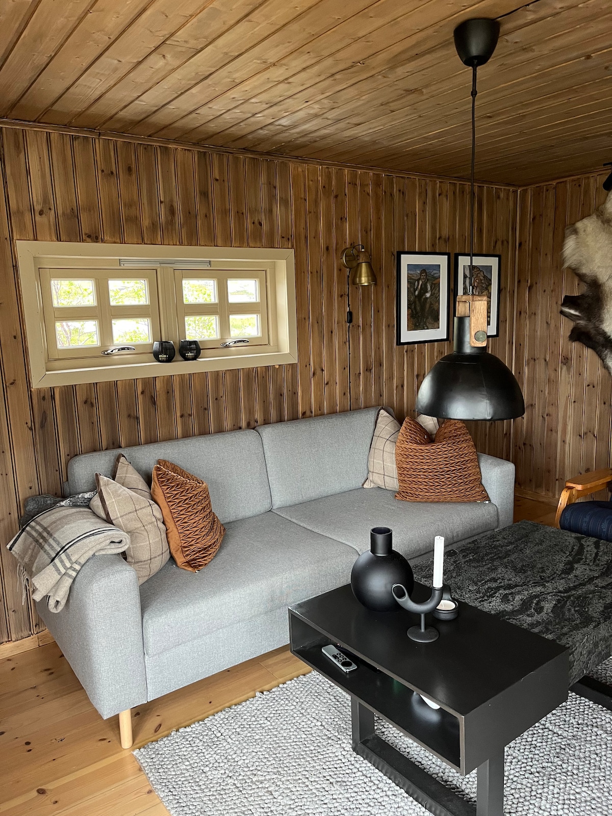 A comfortable seating area is present, featuring a light gray sofa adorned with orange accent pillows. A dark coffee table, equipped with a decorative candle and a pitcher, complements the space. Natural wood paneling lines the walls, while a cozy woven rug adds texture.
