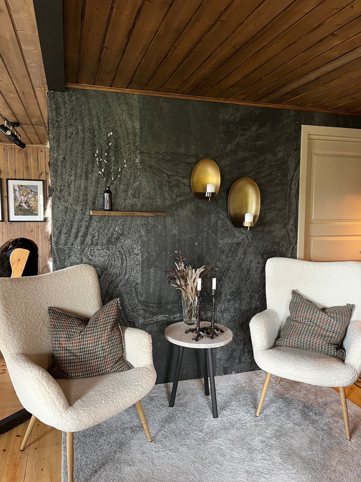 Two armchairs with light upholstery and plaid accent pillows are positioned beside a small round table. The wall behind features a textured dark surface, complemented by two wall-mounted candle holders. Modern decor elements, such as a decorative shelf and dried plants, enhance the inviting space.